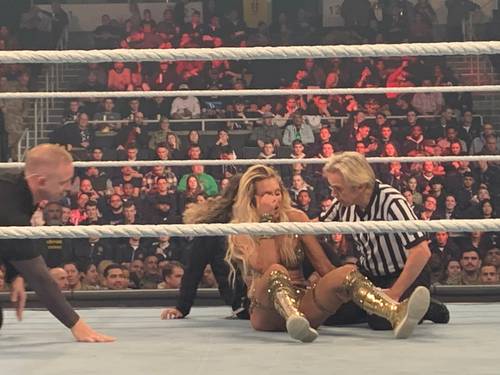 Superluchas - Una mujer, Charlotte Flair, parece haberse lastimado durante un combate de lucha con Asuka, que se lleva a cabo en un ring de lucha libre bajo la supervisión de un árbitro.
