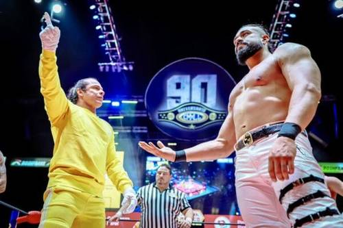 Superluchas - Andrade y Volador Jr., ambos vestidos con trajes amarillos, parados uno al lado del otro.