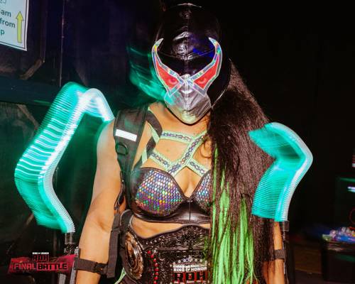 Superluchas - Una mujer vestida con un disfraz sosteniendo una máscara iluminada mientras rinde homenaje a Athena en el evento ROH Final Battle 2023.