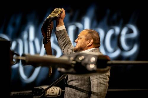 Superluchas - Tony Schiavone, un hombre de traje, sostiene con orgullo un cinturón de lucha libre mientras elogia a Samoa Joe.