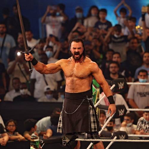 Un hombre con una falda escocesa sosteniendo una espada frente a una multitud durante el momento presente de Drew McIntyre.