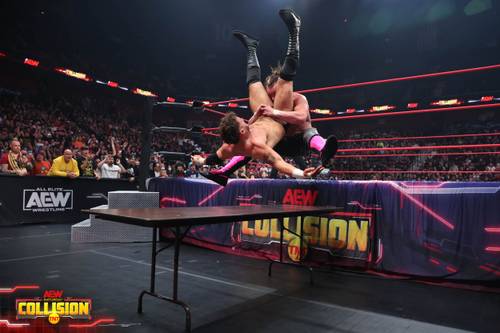 Superluchas - Dos luchadores encima de una mesa en un ring de lucha libre, Bully Ray critica al árbitro de MJF vs. Kenny Omega.