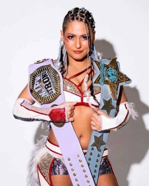 Superluchas - Una mujer vestida de luchadora, Giulia, sosteniendo dos cinturones mientras contempla su futuro en la WWE o el estrellato.